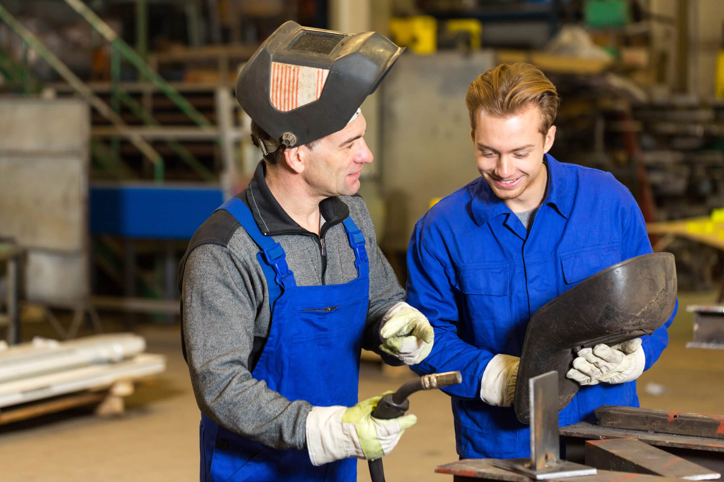 Zwei Handwerker im Blaumann stehen in der Werkstatt und haben ein Schweißgerät in der Hand